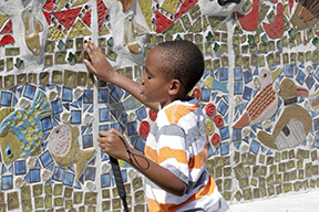 A boy runs his hand along a tile mosaic wall.