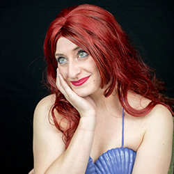 A woman in a red wig and mermaid costume looks up and away while smiling.