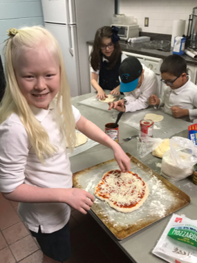 Girl shows off home made pizza while other kids work on their pizzas in the Charlotte SEE Camp kitchen area