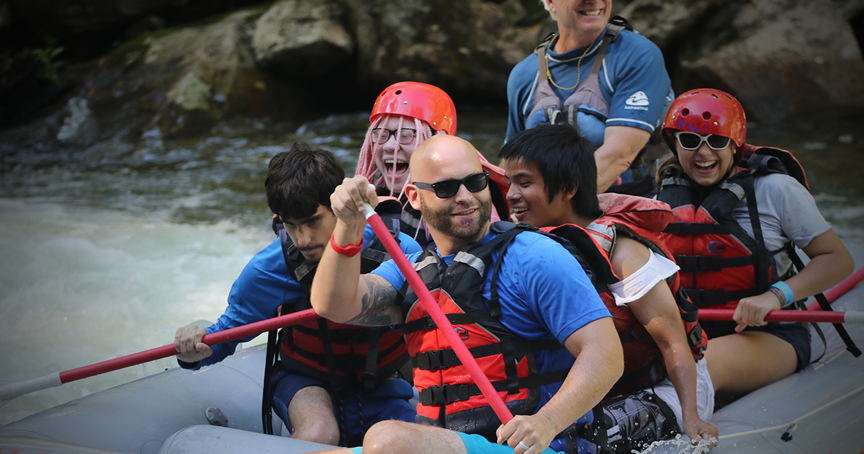 Kids who are blind paddle in a raft down a rapid-filled river lead by a sighted guide