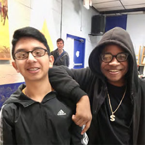two boys stand together, both wearing glasses and smiling