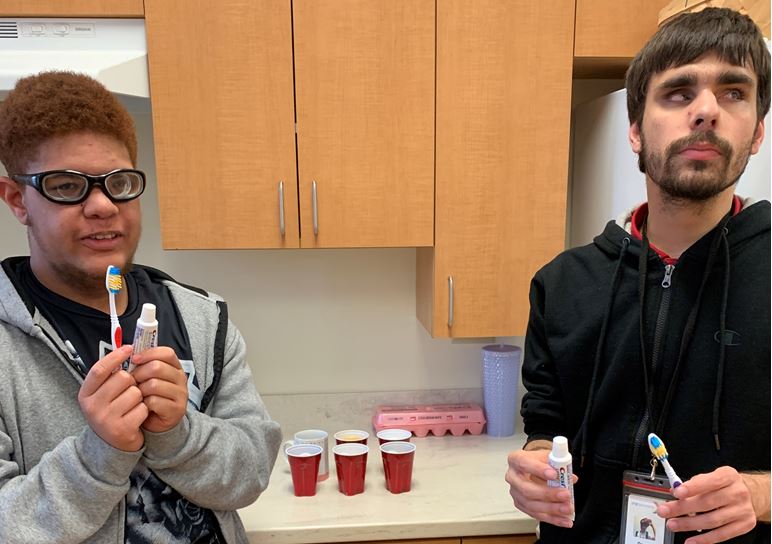 Brandon and Drew with toothbrushes and toothpaste tubes standing in front of a counter with 6 cups of liquid each containing an egg