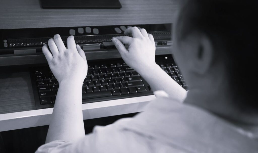 View over shoulder of woman who is blind with hands resting on a braille display reader for computer