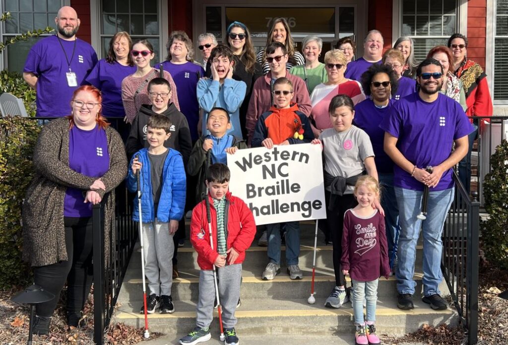 group photo of competitors, staff, volunteers, parents, and teachers on the front steps of Tracy’s Little Red Schoolhouse with a sign that reads Western NC Braille Challenge