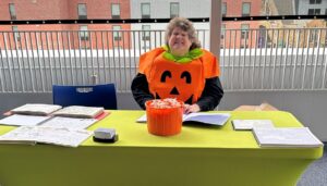 Kim Shoffner dressed as a jack-o-lantern sitting behind a table with Braille books spread out all around and an orange bucket of bubble gum