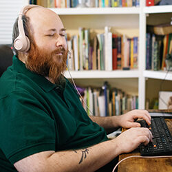 A man wearing headphones sits at a desk with his hands on a keyboard. A bookshelf is visible in the background.