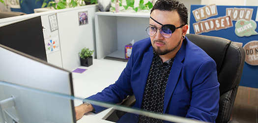 Man with eye patch and wearing glasses sits at desk with hand on the computer mouse