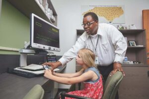 A young girl sitting at a computer, being instructed how to use a screen reader by an adult man.